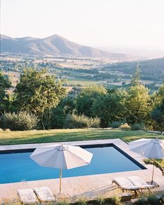 an outdoor swimming pool surrounded by lawn chairs and umbrellas with mountains in the background