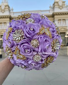 a bridal bouquet with purple roses and gold brooches in front of a building