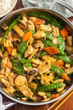 a pan filled with chicken and veggies on top of a wooden table next to rice