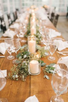 a long wooden table with candles and greenery on it, surrounded by wine glasses