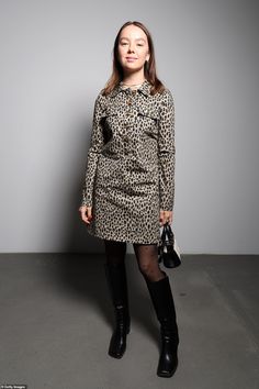 a woman standing in front of a gray wall wearing black boots and a leopard print dress