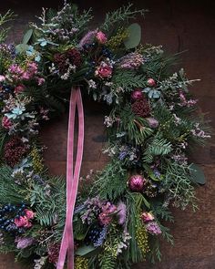 a christmas wreath with pink ribbon hanging on a wooden table, decorated with greenery and berries