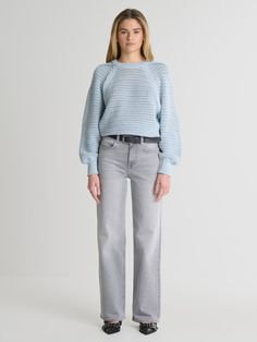 a woman standing in front of a white background wearing grey jeans and a blue sweater