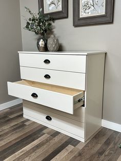 a white dresser with three drawers in front of two pictures on the wall above it