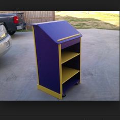 a purple and yellow book shelf sitting in the middle of a driveway next to a car