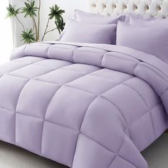 a bed with purple comforter and pillows on it in a white room next to a potted plant