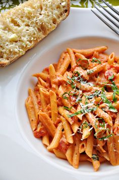 a white plate topped with pasta covered in sauce and parmesan cheese next to a slice of bread