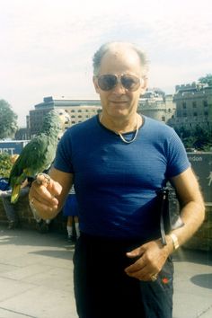 a man holding a green bird on his arm in front of some buildings and trees