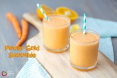 two glasses filled with orange smoothie sitting on top of a cutting board next to sliced carrots