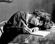 a woman laying down with her head on the desk and writing in a book while holding a pen