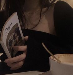 a woman reading a book while sitting at a table with a coffee cup and spoon