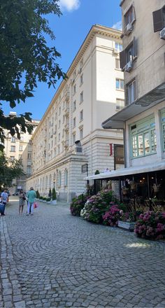 people are walking down the cobblestone street in front of an old building with many windows