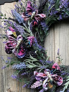 a wreath with purple flowers and greenery hanging on a wooden door, ready to be used as a decoration