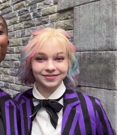 two young women standing next to each other in front of a brick wall wearing purple and black outfits