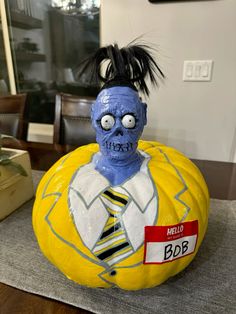 a yellow pumpkin with a zombie head and tie painted on it's face, sitting on a table