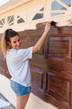 a woman is painting the side of a garage door with brown paint and she is smiling