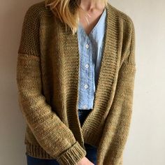 a woman standing in front of a white wall wearing a brown cardigan sweater and jeans