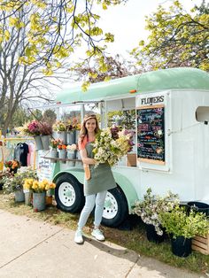 a woman standing in front of a food truck with flowers on the side and potted plants behind her