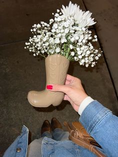 a woman's hand holding a boot shaped vase with flowers in it and her feet on the ground