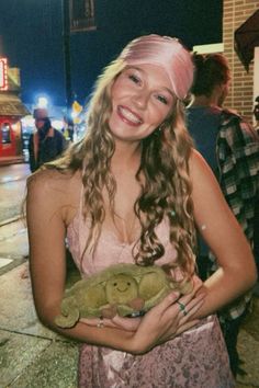 a woman holding a stuffed animal in her hands on the sidewalk at night time with other people standing around