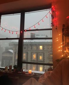 a window with christmas lights hanging from it's sides and the city in the background