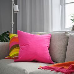 a pink pillow sitting on top of a gray couch next to a white lamp and window