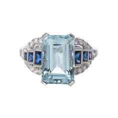 This ring has a vintage-inspired look featuring an emerald-cut aquamarine center flanked by baguette-cut blue sapphires and round brilliant diamond accents set in 18K white gold with milgrain edges. Total aquamarine weight: 4.22ct Total blue sapphire weight: 0.50ct Total diamond weight: 0.31ct Ring size: 7.5 Blue Sapphire Multi-stone Baguette Ring, Blue Multi-stone Sapphire Ring In Art Deco Style, Blue Sapphire Ring With Multi-stone Baguette Cut, Blue Multi-stone Baguette Cut Sapphire Ring, Timeless Blue Multi-stone Sapphire Ring, Blue Aquamarine Baguette-cut Rings, Blue Platinum Rings With Baguette Diamonds, Luxury Blue Baguette Cut Topaz Ring, Blue Formal Rings With Side Stones