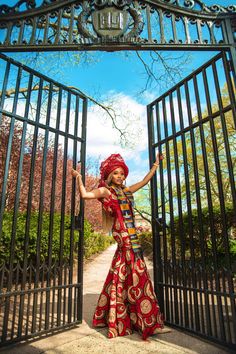 a woman in a red and gold dress is standing at an iron gate with her arms outstretched