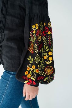 a woman wearing jeans and a black blouse with colorful flowers on the side, holding onto her purse