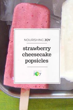 two popsicles in a metal container on a green surface with a white sign that says, nourishing joy strawberry cheesecake pops