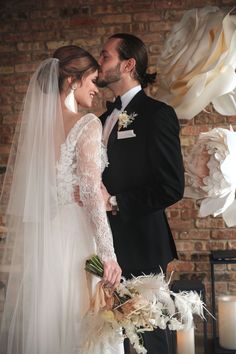 a bride and groom standing next to each other in front of a brick wall with white flowers