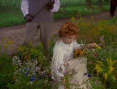 a woman sitting on the ground surrounded by wildflowers and other flowers with a man standing behind her