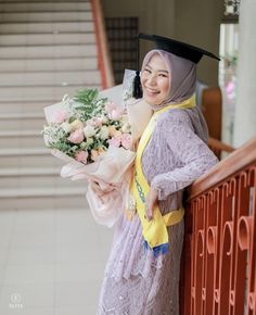 a woman in a graduation gown is holding flowers and smiling at the camera while leaning on a railing