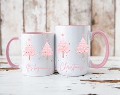 two coffee mugs with christmas trees painted on the sides and pink handles, sitting next to each other