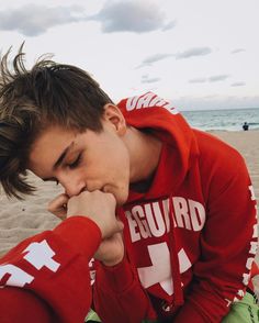 a young man sitting on top of a sandy beach next to the ocean wearing a red hoodie