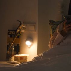 a person sitting on a bed looking at their cell phone in the dark, with a light turned on