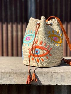 Embrace the mystical allure of the Evil Eye with this uniquely crafted embroidered straw backpack. Handcrafted by skilled Mexican artisans, this boho straw bag large features colorful Evil Eye motifs that add a touch of whimsical charm to your ensemble. The bucket design offers ample space for all your essentials, making it an ideal companion for any adventure or festival outing. This bohemian straw bag exudes a vibrant and playful energy, perfect for adding a pop of color and culture to your lo Embroidered Natural Straw Travel Bag, Embroidered Natural Bags For Vacation, Embroidered Straw Tote Bag For Travel, Bohemian Bucket Bag For Vacation, Summer Festival Bucket Shoulder Bag, Embroidered Beige Straw Bag For Travel, Bohemian Bucket Beach Bag, Bohemian Embroidered Straw Tote Bag, Bohemian Natural Embroidered Straw Bag