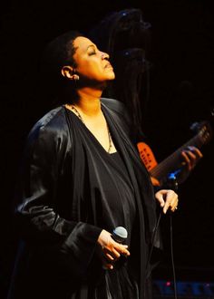 a woman singing into a microphone while holding a guitar in her right hand and wearing a black jacket