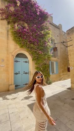 a woman standing in front of a building with purple flowers growing on the side of it