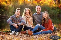 three people sitting on the ground in front of a pond and trees with leaves all around them