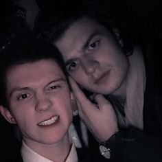 two young men posing for the camera in front of a dark background, one is wearing a suit and the other has his hand on his chin