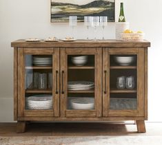 a wooden cabinet with glass doors and plates on the bottom, next to a painting