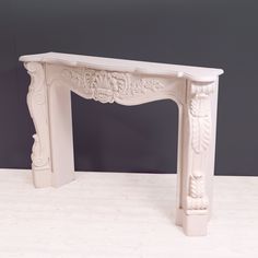 a white marble fireplace surround with carvings on the top and sides, against a gray wall