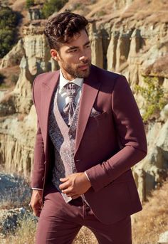 a man wearing a suit and tie standing in front of some rocks with his hands in his pockets