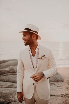 a man in a white suit and hat standing on the rocks by the ocean with his hands in his pockets