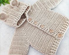 two crocheted sweaters with buttons are shown on a table next to green leaves