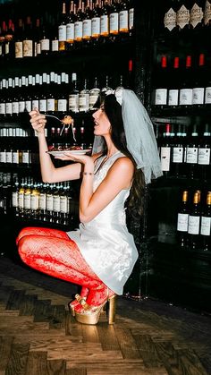 a woman dressed in red and white is sitting on a stool with wine bottles behind her