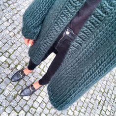 a woman in black pants and green sweater standing on cobblestone street with her legs crossed