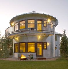 a round house with an outdoor fire pit in the middle of it at dusk, surrounded by trees and grass