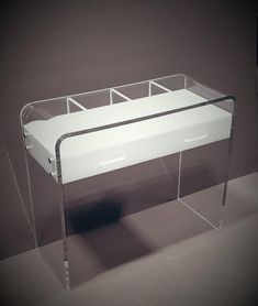 a white desk with clear legs and drawers on it's sides, in front of a gray wall